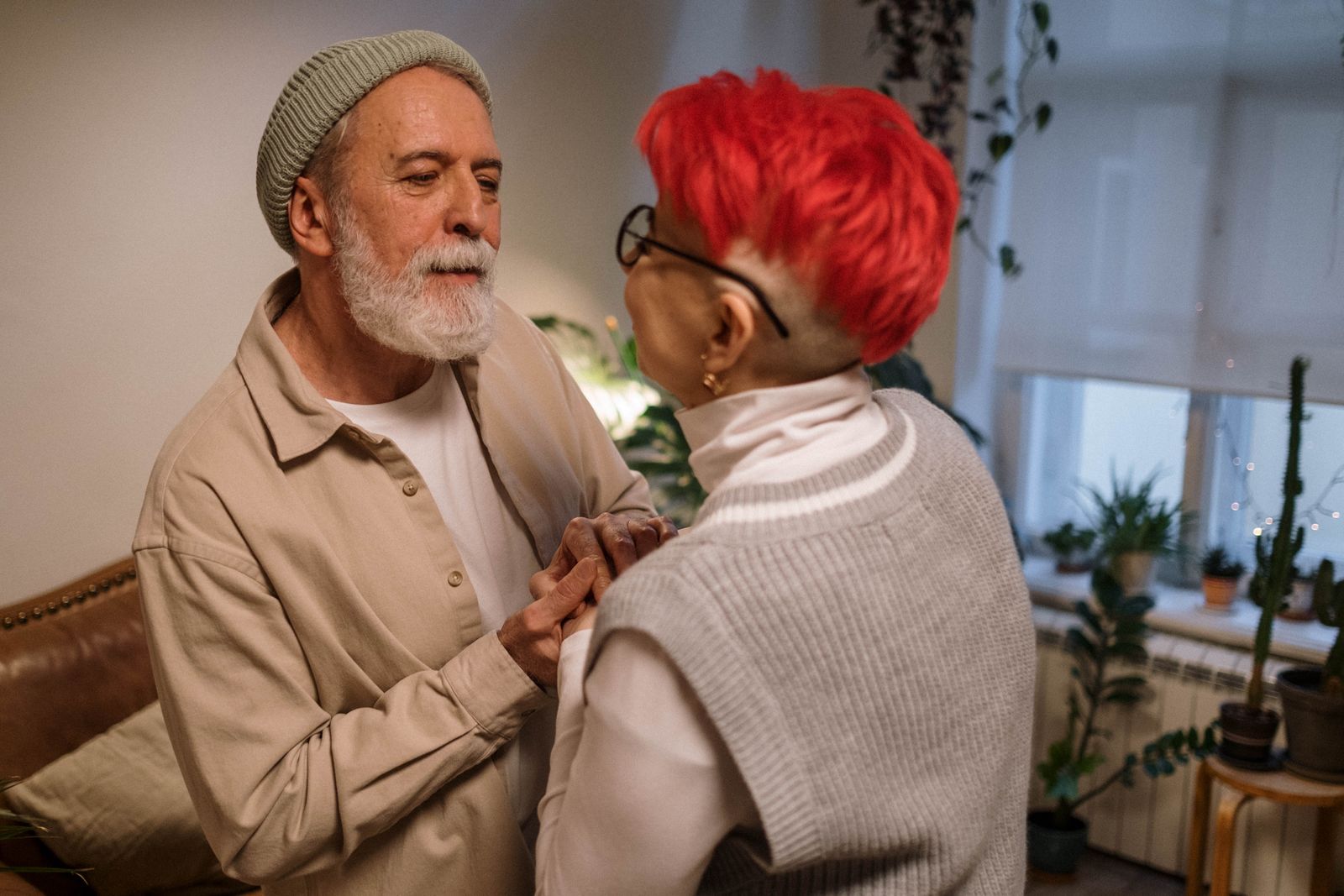An older woman and her caregiver standing close together at home among houseplants, smiling warmly at each other in soft daylight